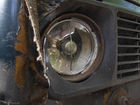 A close-up view of an old, rusted car headlight with visible corrosion around the metal frame. The glass and reflector inside the headlight appear worn, and there is a rough, weathered rope partially in view.