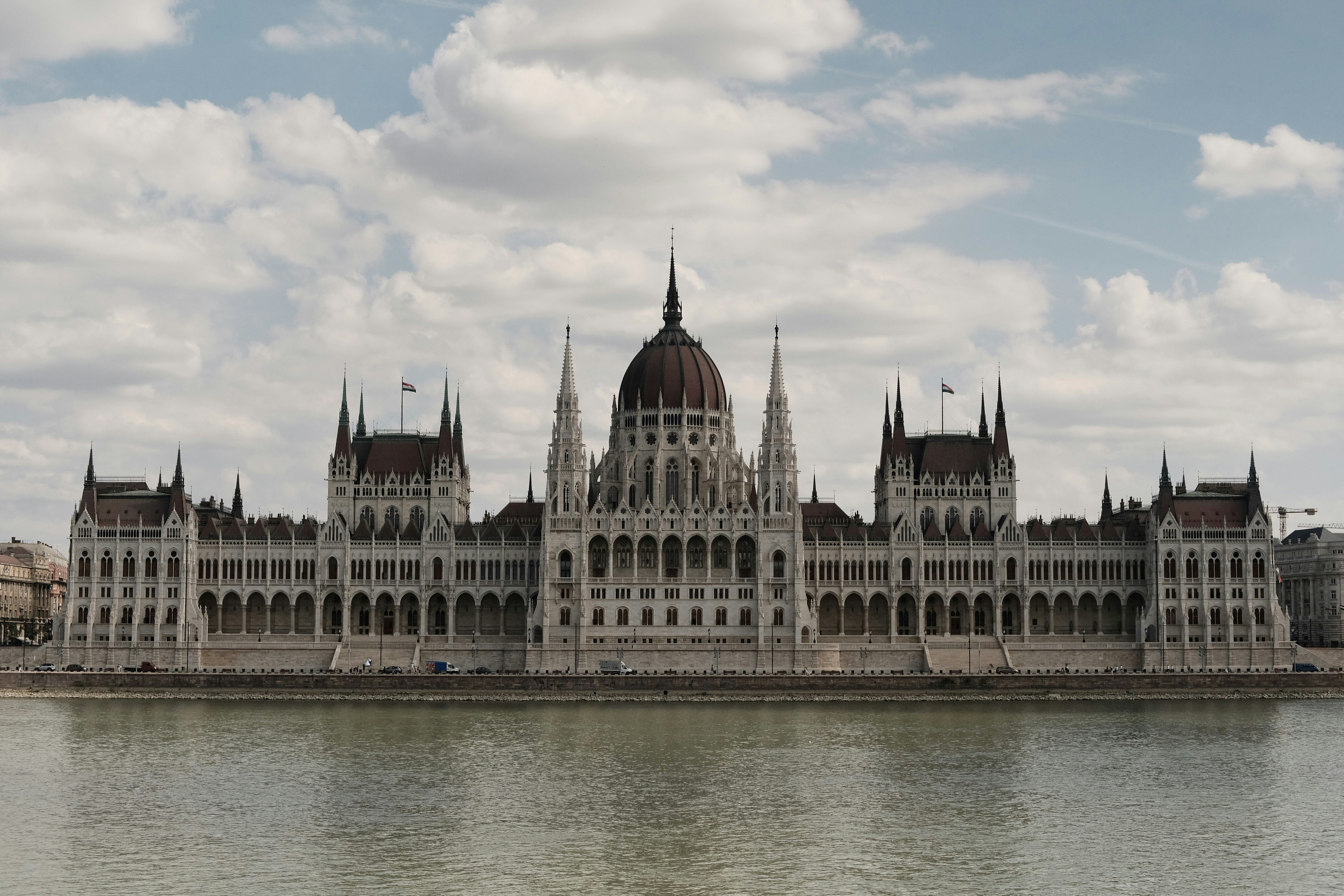a large white building with a domed roof by a body of water with Hungarian Parliament Building in the background
