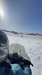 A person wearing a helmet and blue jacket rides a snowmobile across a vast snowy landscape under a bright blue sky. Tracks in the snow indicate previous passage, while the sun shines brightly to the left, casting light across the scene.