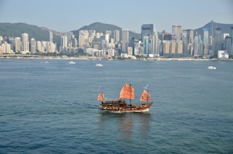 A traditional Asian junk boat with red sails is sailing on a large body of water. In the background, a densely packed urban skyline with high-rise buildings and mountains beyond the city.