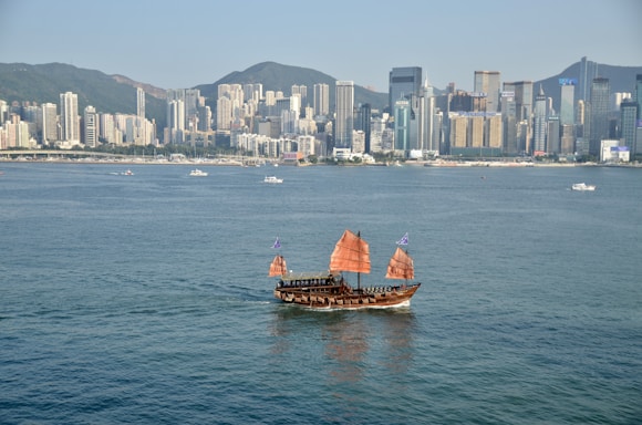 A traditional Asian junk boat with red sails is sailing on a large body of water. In the background, a densely packed urban skyline with high-rise buildings and mountains beyond the city.
