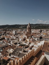 An aerial view of a Spanish town showcasing a dense assembly of terracotta-roofed buildings and narrow streets. A prominent historical church tower stands out in the foreground, while rugged hills stretch out in the background under a clear blue sky.