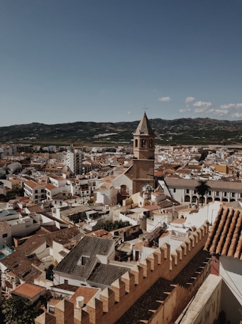 An aerial view of a Spanish town showcasing a dense assembly of terracotta-roofed buildings and narrow streets. A prominent historical church tower stands out in the foreground, while rugged hills stretch out in the background under a clear blue sky.