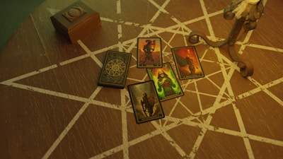 A wooden table covered with intricate white geometric patterns and holding several colorful tarot cards depicting various figures and symbols. There is also a wooden box on the table and a base of a metal decorative object.