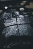 A close-up view of a dark, wet surface with raindrops. The surface appears to be leather or similar material, and it is secured with a net or bungee cord. The droplets create a shiny texture, reflecting light in the dim setting.