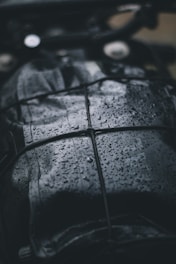 A close-up view of a dark, wet surface with raindrops. The surface appears to be leather or similar material, and it is secured with a net or bungee cord. The droplets create a shiny texture, reflecting light in the dim setting.
