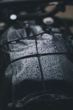 A close-up view of a dark, wet surface with raindrops. The surface appears to be leather or similar material, and it is secured with a net or bungee cord. The droplets create a shiny texture, reflecting light in the dim setting.