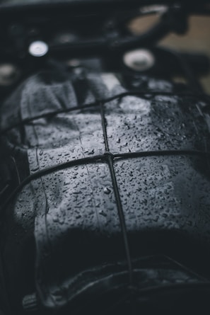 A close-up view of a dark, wet surface with raindrops. The surface appears to be leather or similar material, and it is secured with a net or bungee cord. The droplets create a shiny texture, reflecting light in the dim setting.