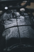 A close-up view of a dark, wet surface with raindrops. The surface appears to be leather or similar material, and it is secured with a net or bungee cord. The droplets create a shiny texture, reflecting light in the dim setting.