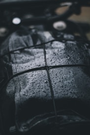 A close-up view of a dark, wet surface with raindrops. The surface appears to be leather or similar material, and it is secured with a net or bungee cord. The droplets create a shiny texture, reflecting light in the dim setting.