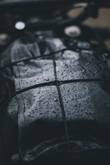 A close-up view of a dark, wet surface with raindrops. The surface appears to be leather or similar material, and it is secured with a net or bungee cord. The droplets create a shiny texture, reflecting light in the dim setting.