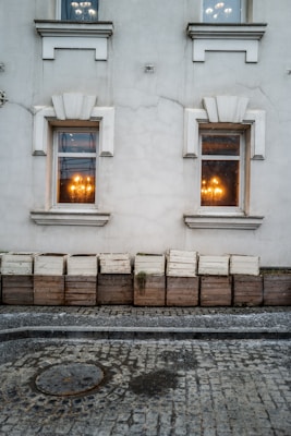 Two windows on a weathered building wall, each illuminated by a warm glowing chandelier visible through the glass. Below the windows is a row of wooden planters, some with sparse vegetation. The ground is cobblestone with a circular metal manhole cover.
