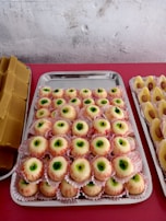 A table displaying trays of sweet treats, including a batch of confectionery resembling eyeballs with a white outer layer, green center, and red detailing. Flanking these are other sweets with nut toppings on either side, and a stack of rectangular brown candies on the left.