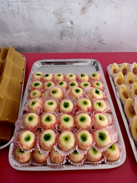 A table displaying trays of sweet treats, including a batch of confectionery resembling eyeballs with a white outer layer, green center, and red detailing. Flanking these are other sweets with nut toppings on either side, and a stack of rectangular brown candies on the left.