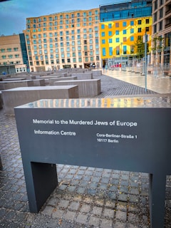 A memorial site featuring several concrete slabs of varying heights arranged in a grid pattern. Nearby are modern buildings with a distinct yellow-painted facade. The nearest slab has an inscription about the Memorial to the Murdered Jews of Europe in Berlin.