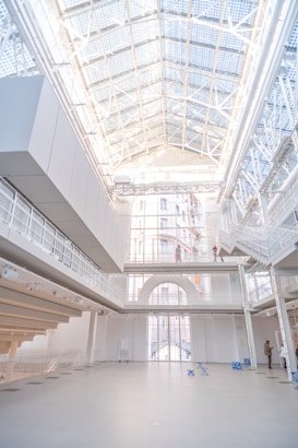 A spacious, modern interior with a high, glass-paneled ceiling allowing natural light to flood in. The atrium is dominated by white, minimalist architecture with metal beams and railings. A few people are visible in the background, standing on different levels. Blue folding chairs are scattered on the floor.