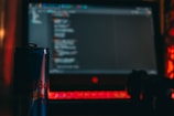 A dark room with a laptop displaying code on the screen. In the foreground, there is a can of energy drink and a game controller, all illuminated by the red glow of the laptop's keyboard.