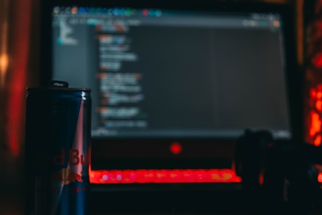 A dark room with a laptop displaying code on the screen. In the foreground, there is a can of energy drink and a game controller, all illuminated by the red glow of the laptop's keyboard.