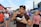 A person enjoys food at an outdoor event, holding a chicken wing and a bottle of water. Surrounding them are various people, vendor stalls, and a stadium in the background. The atmosphere suggests a lively gathering, possibly a festival or fair.