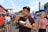 A person enjoys food at an outdoor event, holding a chicken wing and a bottle of water. Surrounding them are various people, vendor stalls, and a stadium in the background. The atmosphere suggests a lively gathering, possibly a festival or fair.