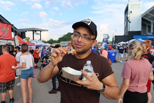 A person enjoys food at an outdoor event, holding a chicken wing and a bottle of water. Surrounding them are various people, vendor stalls, and a stadium in the background. The atmosphere suggests a lively gathering, possibly a festival or fair.
