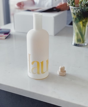 A frosted glass bottle with minimalist design featuring light yellow text sits on a white marble countertop. The bottle is accompanied by a matching cylindrical cap placed beside it. In the background, a person is handling a magazine or catalog, and there is a vase with green leafy plants.