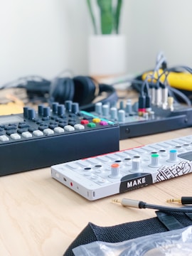 A tabletop with electronic music equipment, including a synthesizer with colorful knobs and buttons, and a mixer featuring numerous controls. Headphones and various cables are also present, with a potted plant in the background.