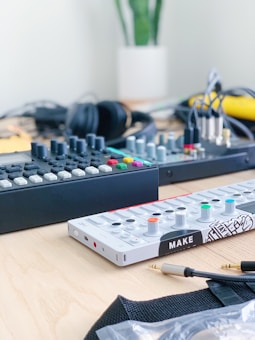 A tabletop with electronic music equipment, including a synthesizer with colorful knobs and buttons, and a mixer featuring numerous controls. Headphones and various cables are also present, with a potted plant in the background.