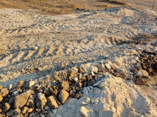 Tire tracks are imprinted in sandy and rocky terrain. The ground is uneven, with scattered rocks and small mounds of dirt. The area appears dry and barren, typical of a construction or off-road area.