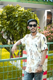 A young man is leaning against a metal fence in a garden setting. He is wearing a floral-patterned shirt, sunglasses, and a watch. Behind him, there is dense foliage and a sign written in Bengali, suggesting the location might be near a public place, possibly a park or an institution in Bangladesh.