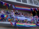 A group of people stand on a balcony decorated with colorful Pride flags and banners. The decorations include rainbow patterns and messages promoting love and gender inclusivity. Some people are leaning on the railing, engaging in conversation, and wearing accessories that match the vibrant theme.
