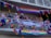 A group of people stand on a balcony decorated with colorful Pride flags and banners. The decorations include rainbow patterns and messages promoting love and gender inclusivity. Some people are leaning on the railing, engaging in conversation, and wearing accessories that match the vibrant theme.