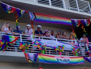 A group of people stand on a balcony decorated with colorful Pride flags and banners. The decorations include rainbow patterns and messages promoting love and gender inclusivity. Some people are leaning on the railing, engaging in conversation, and wearing accessories that match the vibrant theme.