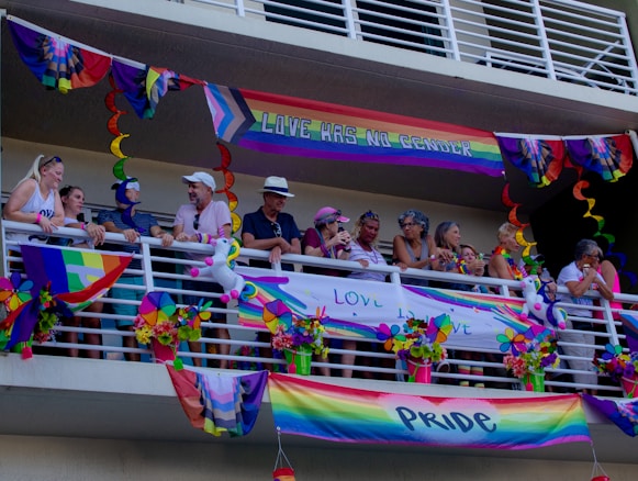 A group of people stand on a balcony decorated with colorful Pride flags and banners. The decorations include rainbow patterns and messages promoting love and gender inclusivity. Some people are leaning on the railing, engaging in conversation, and wearing accessories that match the vibrant theme.