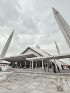 A large, modern mosque with angular architectural design, featuring tall minarets reaching upwards towards a cloudy sky. People are seen walking up the wide steps leading to the entrance, dressed in various colors.