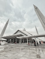 A large, modern mosque with angular architectural design, featuring tall minarets reaching upwards towards a cloudy sky. People are seen walking up the wide steps leading to the entrance, dressed in various colors.