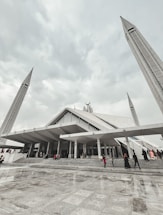A large, modern mosque with angular architectural design, featuring tall minarets reaching upwards towards a cloudy sky. People are seen walking up the wide steps leading to the entrance, dressed in various colors.