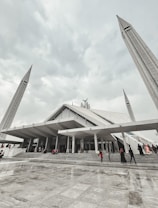A large, modern mosque with angular architectural design, featuring tall minarets reaching upwards towards a cloudy sky. People are seen walking up the wide steps leading to the entrance, dressed in various colors.