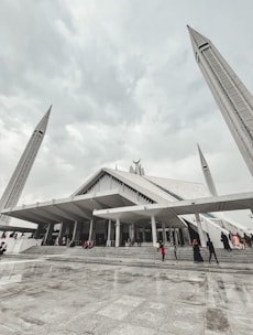 A large, modern mosque with angular architectural design, featuring tall minarets reaching upwards towards a cloudy sky. People are seen walking up the wide steps leading to the entrance, dressed in various colors.