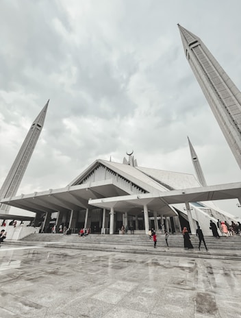 A large, modern mosque with angular architectural design, featuring tall minarets reaching upwards towards a cloudy sky. People are seen walking up the wide steps leading to the entrance, dressed in various colors.