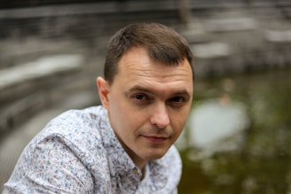 A man with short dark hair is wearing a white shirt with blue floral patterns. The background features a blurred outdoor setting with a stone structure and water, giving an impression of an urban location.