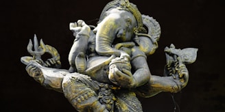 An intricately detailed statue of Ganesha, the elephant-headed Hindu deity, depicted with multiple arms holding various symbolic objects. The statue features ornate carvings and a realistic texture, with a patina of age visible on the surface.