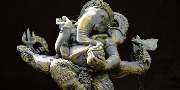 An intricately detailed statue of Ganesha, the elephant-headed Hindu deity, depicted with multiple arms holding various symbolic objects. The statue features ornate carvings and a realistic texture, with a patina of age visible on the surface.