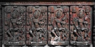 A row of intricately carved stone reliefs depicting mythological figures, possibly deities, set against a weathered stone background. The carvings exhibit detailed artistry with figures in various poses, adorned with ornate headgear and garments.