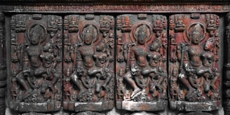 A row of intricately carved stone reliefs depicting mythological figures, possibly deities, set against a weathered stone background. The carvings exhibit detailed artistry with figures in various poses, adorned with ornate headgear and garments.