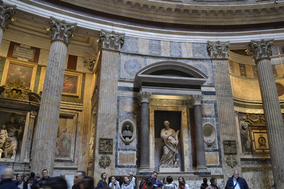 The image depicts a large, ornate room with marble columns and walls adorned with paintings. The ceiling features a circular design that adds architectural interest to the space. A statue is prominently displayed on one side of the room, surrounded by several other statues. People can be seen walking around the room, adding a sense of scale and activity to the scene.