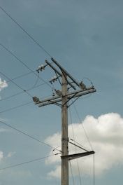 A tall utility pole stands against a clear blue sky, with electrical wires stretching across it. The pole is made of wood, and several insulators and transformer components can be seen attached to it. Puffy white clouds float in the background, adding a serene contrast to the industrial structure.