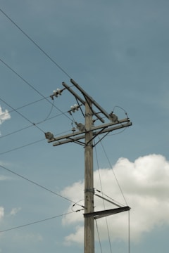 A tall utility pole stands against a clear blue sky, with electrical wires stretching across it. The pole is made of wood, and several insulators and transformer components can be seen attached to it. Puffy white clouds float in the background, adding a serene contrast to the industrial structure.