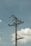 A tall utility pole stands against a clear blue sky, with electrical wires stretching across it. The pole is made of wood, and several insulators and transformer components can be seen attached to it. Puffy white clouds float in the background, adding a serene contrast to the industrial structure.