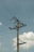A tall utility pole stands against a clear blue sky, with electrical wires stretching across it. The pole is made of wood, and several insulators and transformer components can be seen attached to it. Puffy white clouds float in the background, adding a serene contrast to the industrial structure.